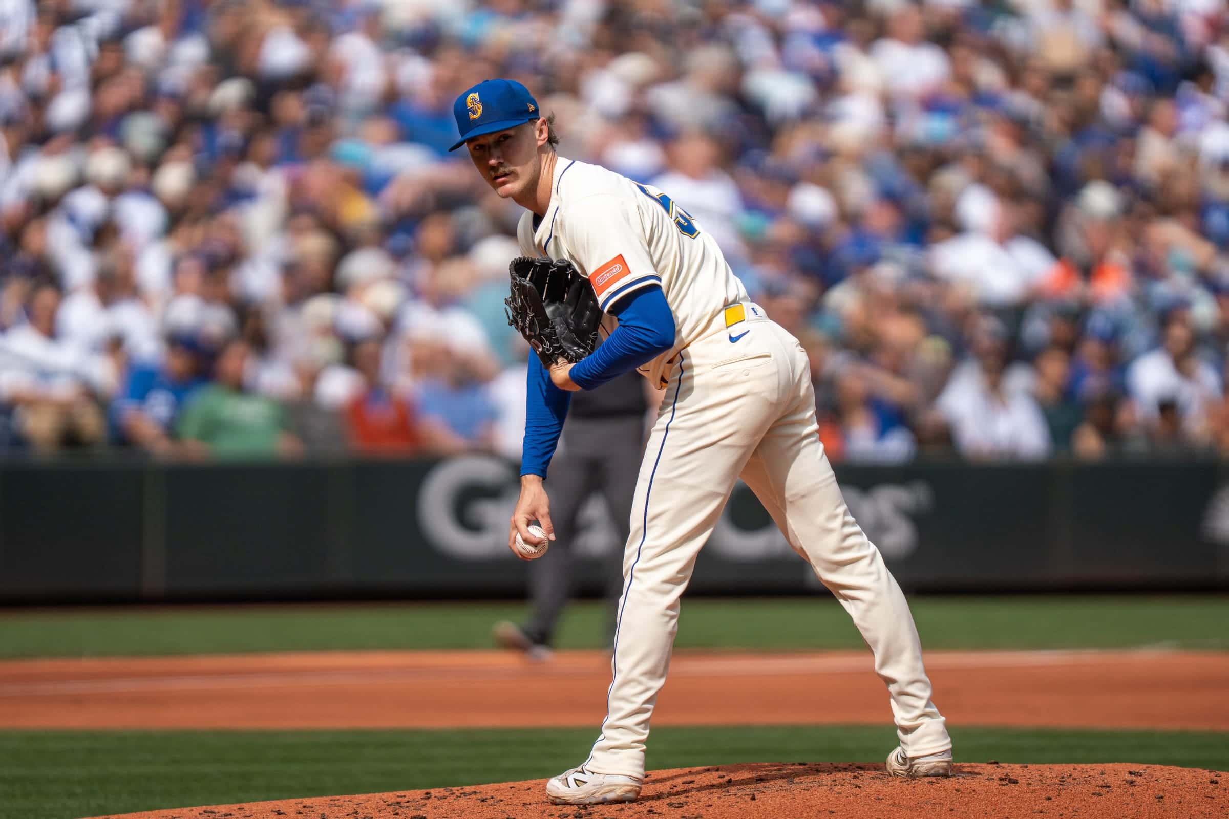 Mariners pitcher in a cream uniform and blue cap looking in toward the plate with the ball at his side