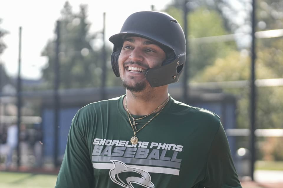 Shoreline baseball player grinning in a batting helmet and a green Shoreline Phins Baseball shirt at practice